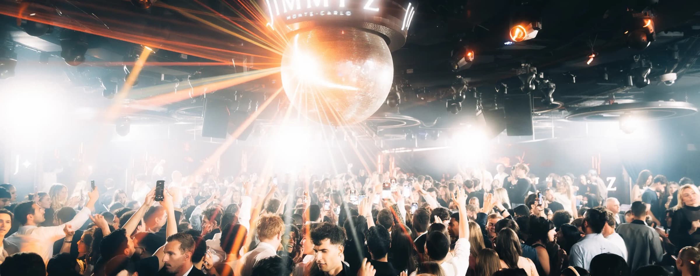 La piste de Jimmy'z Monte-Carlo · foule sous une immense boule à facettes, faisceaux de lumière et enseigne lumineuse