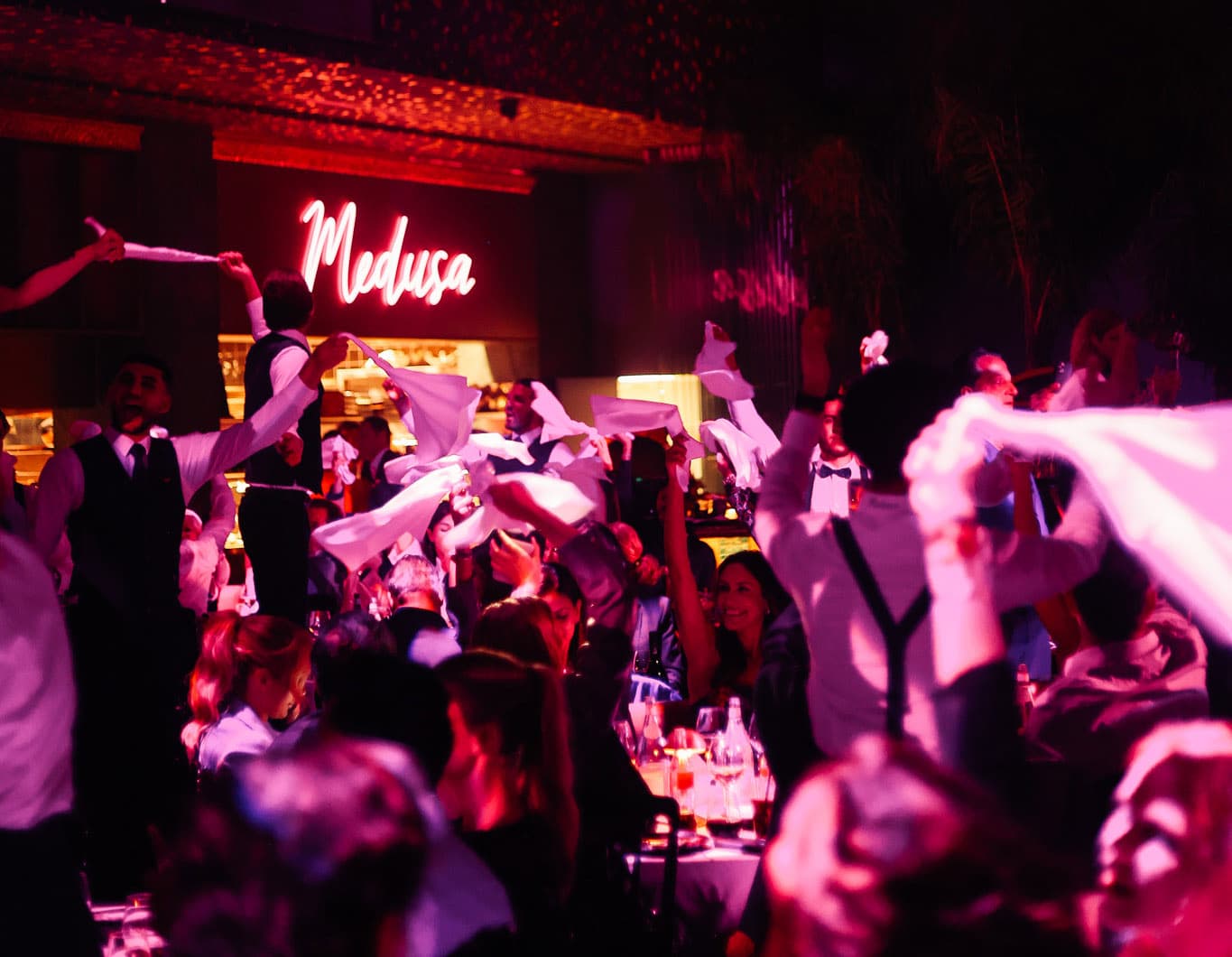L'enseigne néon Medusa au-dessus d'une salle comble, serveurs en gilet agitant des serviettes blanches sous une lumière rose.